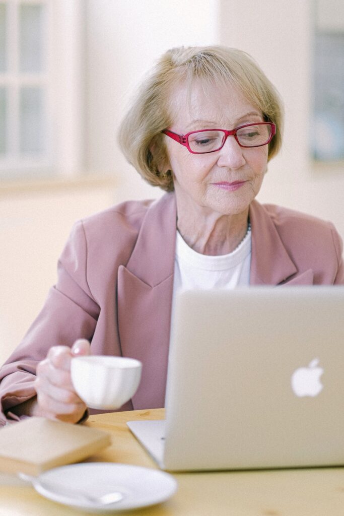 Mature woman in eyeglasses with cup checking important documents on netbook on blurred background